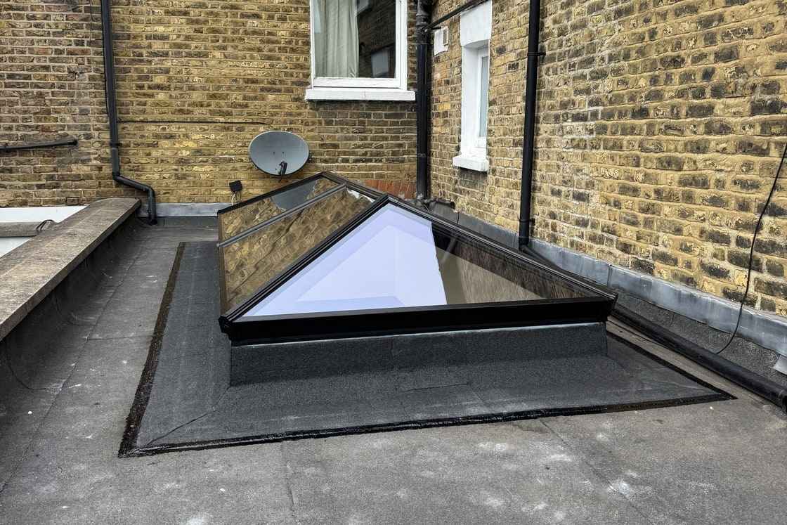 Glazed roof lantern seated on a felt-dressed kerb in a flat felt roof, with the cap sheet apron carried up the upstand and closed under the frame.